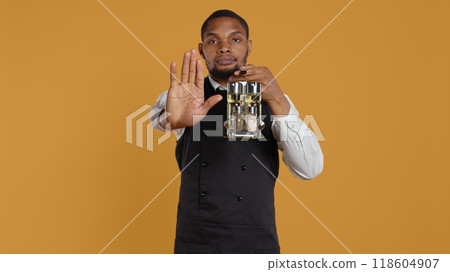 Waiter in uniform showing a stop sign with his palm raised in studio, expressing negative symbol and refusal about something. Showing rejection and being dissatisfied. Camera B. 118604907