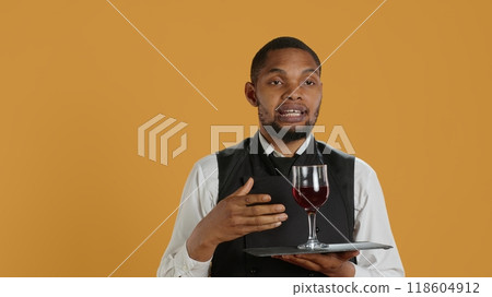 Waiter serving clients with a glass of red wine before dinner, recommending a special drink for customer satisfaction. Employee carrying a tray and serves people with professionalism. Camera A. Waiter serving clients with a glass of red wine before dinner, recommending a special drink for customer satisfaction. Employee carrying a tray and serves people with professionalism. Camera A. 118604912