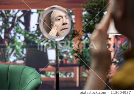 Elderly man looking in the mirror at his wrinkles and aging skin while he does his daily routine for grooming and skincare. Applying rejuvenating cream and lotion for hydration. 118604927