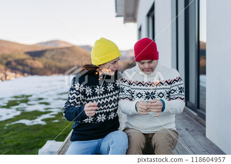 The mother and young man with Down syndrome are outside, having fun with Christmas sparklers and enjoying first snow The mother and young man with Down syndrome are outside, having fun with Christmas sparklers and enjoying first snow 118604957