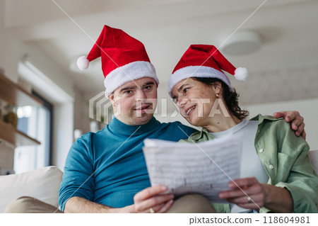 Young man with Down syndrome learning and singing christmas songs with mother. Concept of peaceful Christmas family moment. Young man with Down syndrome learning and singing christmas songs with mother. Concept of peaceful Christmas family moment. 118604981