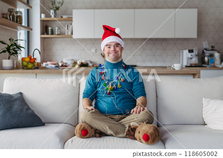 Portrait of young man with Down syndrome with santa's hat on head and christmas light around body, looking at camera. 118605002