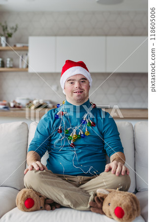 Portrait of young man with Down syndrome with santa's hat on head and christmas light around body, looking at camera. 118605006