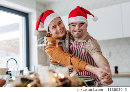 Young man with Down syndrome and his mom dancing while baking Christmas gingerbread cookies in kitchen. Christmas peaceful moment for man with Down syndrome. Young man with Down syndrome and his mom dancing while baking Christmas gingerbread cookies in kitchen. Christmas peaceful moment for man with Down syndrome. 118605043