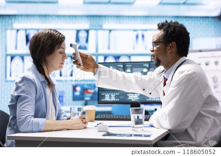 General practitioner using a thermometer to measure fever on a female patient, doing a check up visit examination with modern medical tools. Doctor specialist checking temperature on a woman. 118605052
