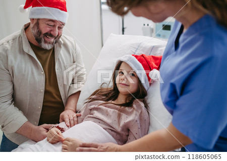 Father beside daughter on hospital bed. Sick little girl spending Christmas in hospital Nurse working shift on Christmas Eve. 118605065