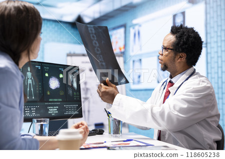 Professional medic analyzing x ray imaging and test findings in office, giving the diagnostic report for female patient at check up. African american specialist examines scans for interpretation. Professional medic analyzing x ray imaging and test findings in office, giving the diagnostic report for female patient at check up. African american specialist examines scans for interpretation. 118605238