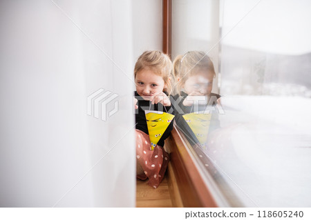 Cute little girl is sitting on indoor windowsill, hiding behind the curtains. 118605240