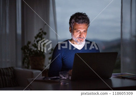 Handsome man working from home office at evening, using laptop. Screen glow illuminates man's face. Telecommuting and home office. 118605309
