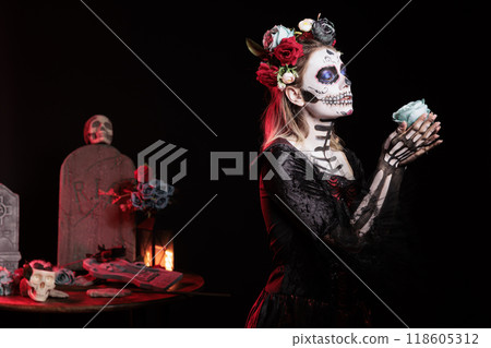 Woman gifting blue rose to dead relatives in graveyard, celebrating Dia de los Muertos. Mysterious lady with flower surrounded by frightening funeral items during Halloween horror event 118605312