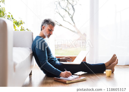Homeoffice for handsome man. Man sitting on floor and working on laptop, writing notes in planner. Telecommuting. Homeoffice for handsome man. Man sitting on floor and working on laptop, writing notes in planner. Telecommuting. 118605317
