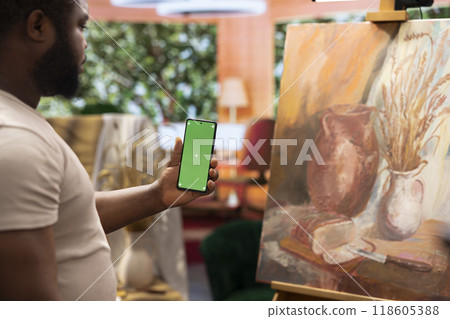African american painter using a gadget with copy space screen in workshop, working on his new nature inspired masterpiece on canvas. Using watercolor paint and creativity in atelier. 118605388