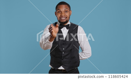 Bellboy hotel porter welcoming guests at the five star lodging resort, smiling and greeting people against blue background. Employee wearing a tuxedo and tie, tourism industry. Camera B. Bellboy hotel porter welcoming guests at the five star lodging resort, smiling and greeting people against blue background. Employee wearing a tuxedo and tie, tourism industry. Camera B. 118605424