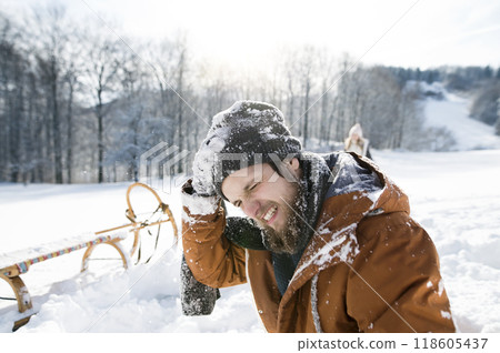 Portrait of man with head injury after snowball fight. Man sitting on ground, snow stuck to clothes and beard. 118605437