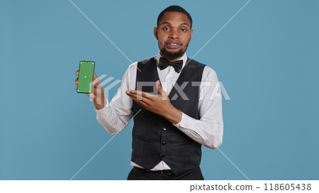 Hotel worker bellhop points at phone with isolated mockup display, showing green screen against blue background in studio. Hotelier doorman greeting resort guests at reception. Camera B. 118605438