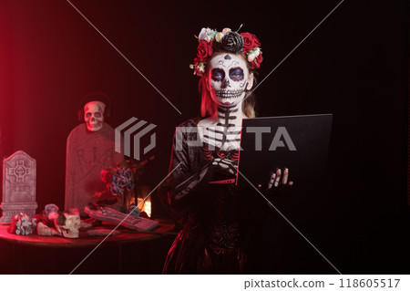 Portrait of woman typing on notebook in graveyard, celebrating Day of the Dead. Mysterious lady working on laptop, surrounded by frightening funeral items during Halloween horror event Portrait of woman typing on notebook in graveyard, celebrating Day of the Dead. Mysterious lady working on laptop, surrounded by frightening funeral items during Halloween horror event 118605517