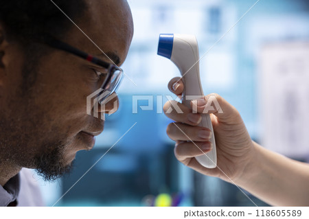 Female doctor taking the temperature of a patient in medical office, checking fever and other flu symptoms at consultation. Physician measuring with a digital thermometer in cabinet. 118605589