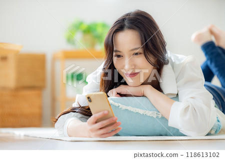 A young woman lying down on a table in the living room and looking at her smartphone 118613102
