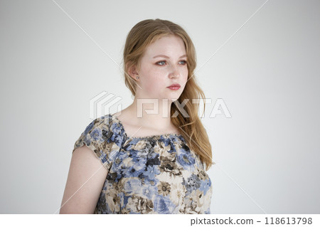 Young woman with long hair poses thoughtfully in a floral blouse against a plain background. The neutral background highlights her thoughtful demeanor. Young adult, european. 118613798