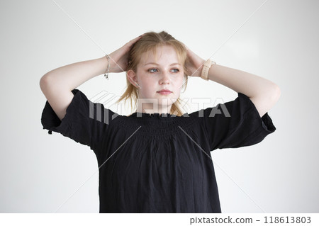 A young woman stands confidently in a minimalist studio, her hands resting on her head as she gazes thoughtfully ahead. The soft lighting highlights her natural beauty. 118613803