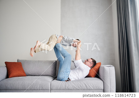 Joyful father lifting happy little son at home. Carefree father and son weekend. Happy young father lying on couch and fooling around with kid. 118614723