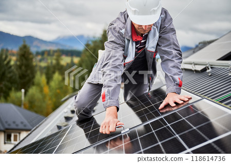 Man engineer mounting photovoltaic solar panels on roof of house. Technician in helmet installing solar module system with help of hex key. Concept of alternative, renewable energy. 118614736