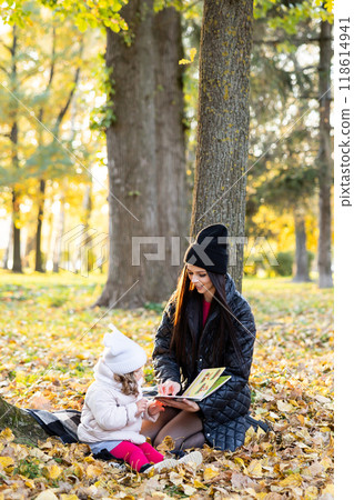 A mother is reading a book with her cute daughter while sitting on the lawn in the park 118614941