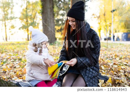 A mother is reading a book with her cute daughter while sitting on the lawn in the park A mother is reading a book with her cute daughter while sitting on the lawn in the park 118614943
