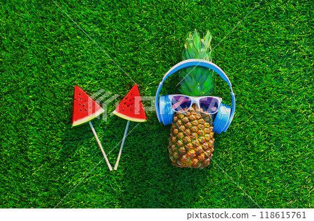 Summer pineapple with headphones listening to music and lollipop or ice cream watermelon on grass Summer pineapple with headphones listening to music and lollipop or ice cream watermelon on grass 118615761