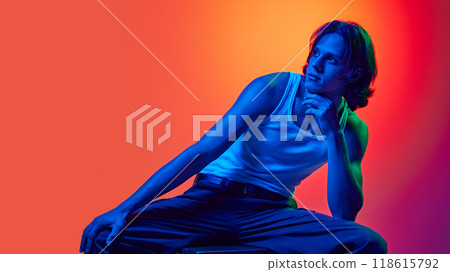 fashion portrait of young attractive man posing sitting on wooden stool dressed casual outfit in blue neon light against gradient studio background. 118615792