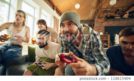 Cheers as two of guys playing football on console, laughter filling cozy room. Popcorn, chips and alcohol drinks keep group energized for their intense matches. Cheers as two of guys playing football on console, laughter filling cozy room. Popcorn, chips and alcohol drinks keep group energized for their intense matches. 118615904
