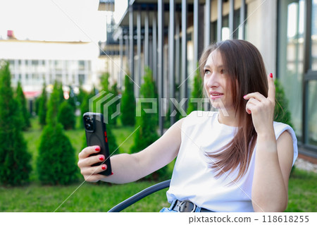 young woman takes selfie makes video call while sitting outdoors, gesturing with her hand. young woman takes selfie makes video call while sitting outdoors, gesturing with her hand. 118618255
