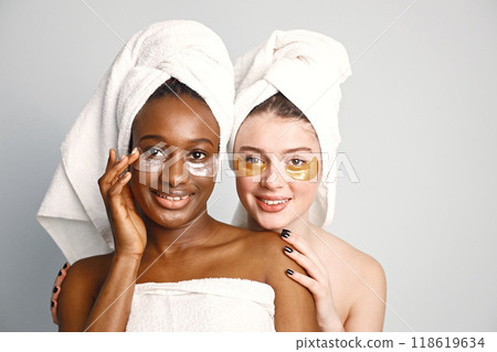 Two young women in white bathrobes standing isolated on white. Girls wearing white towels on their hair. Girls have a eye patches on their faces. 118619634