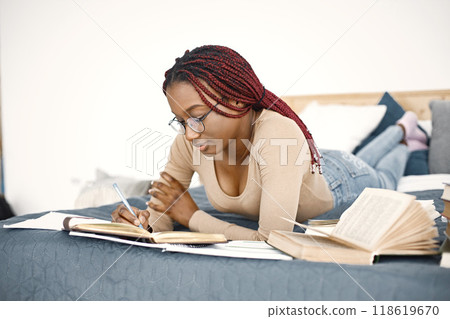 Young black woman lying down on bed reading a book at home. Girl wearing eyeglasses and beige sweater. Girl preparing her homework. Young black woman lying down on bed reading a book at home. Girl wearing eyeglasses and beige sweater. Girl preparing her homework. 118619670