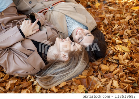 Two girls lying on the yellow leaves at the autumn park. Portrait of female friends. Two girls wearing beige coats. Two girls lying on the yellow leaves at the autumn park. Portrait of female friends. Two girls wearing beige coats. 118619682