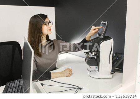 Female optician using laptop at her working place. Woman wearing eyeglasses in ophthalmic shop. Optician checking eyeglasses in special machine Female optician using laptop at her working place. Woman wearing eyeglasses in ophthalmic shop. Optician checking eyeglasses in special machine 118619714