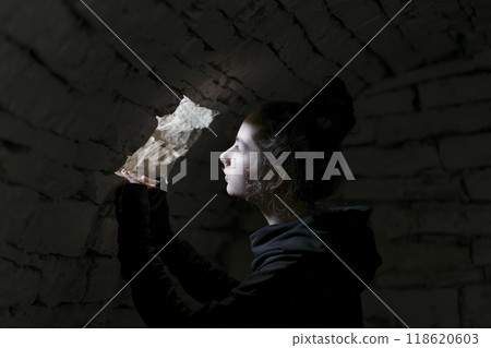 Young adult in a basement looking in a small window. A beam of light falls on her face. Portrait of the young girl in the dungeon with dark background. 118620603