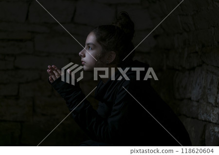 Girl in a bomb shelter prays to God for peace in her country. Portrait of the young woman in the dungeon with dark background. Young adult's face is illuminated by light coming from the opening. Girl in a bomb shelter prays to God for peace in her country. Portrait of the young woman in the dungeon with dark background. Young adult's face is illuminated by light coming from the opening. 118620604