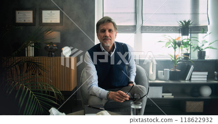 Professional mature therapist or psychologist sits in chair, smiles and looks at camera Professional mature therapist or psychologist sits in chair, smiles and looks at camera 118622932