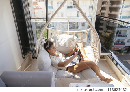A woman is relaxing in a hammock on her balcony, enjoying the warm sunshine on a beautiful day A woman is relaxing in a hammock on her balcony, enjoying the warm sunshine on a beautiful day 118625482