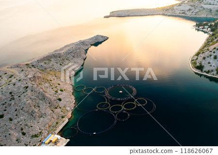 Fish farm operations are visible near the rocky coast as the sun sets in the background. Fish farm operations are visible near the rocky coast as the sun sets in the background. 118626067