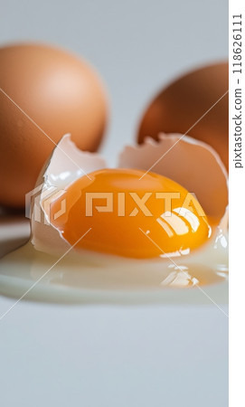 A cracked eggshell sits on a smooth surface, with a bright yellow yolk and clear egg white spilling out. Two whole eggs are in the background, emphasizing freshness and natural textures 118626111