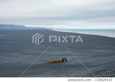 A lone tractor and trailer on a vast black sand beach in Iceland. The beach meets the ocean under an overcast sky, with distant mountains along the coastline. 118626510