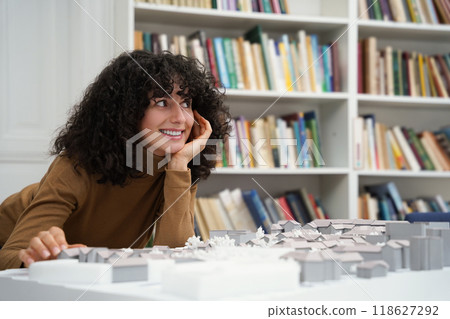 Curly-haired young woman sitting at the table and working on a layout Curly-haired young woman sitting at the table and working on a layout 118627292