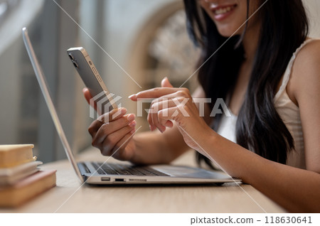 A woman using her smartphone while sitting at a table in a cafe with her laptop placed on the table. A woman using her smartphone while sitting at a table in a cafe with her laptop placed on the table. 118630641