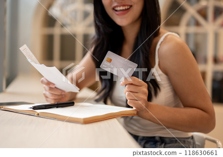 A close-up of a happy Asian woman holding a credit card and reviewing receipts at a table indoors. A close-up of a happy Asian woman holding a credit card and reviewing receipts at a table indoors. 118630661