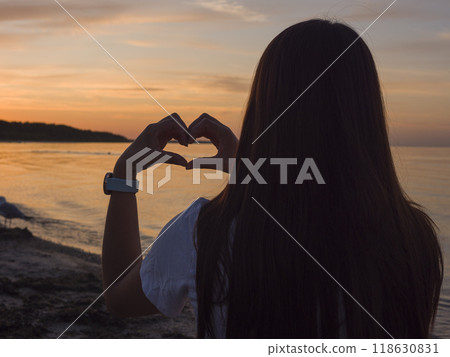 A young woman forms heart with her hands at sunset by seashore warm glow of the sun reflecting her heart's expression. Her silhouette and heart shape are beautifully framed against the vibrant sky. 118630831