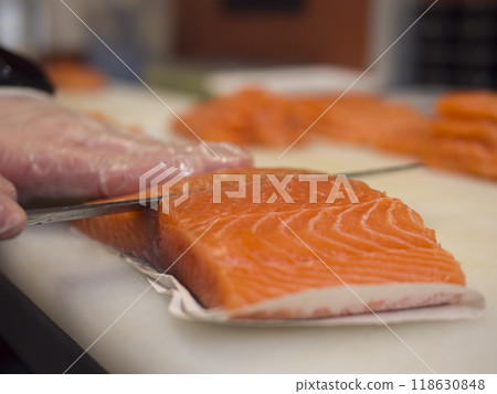 A skilled chef slices fresh salmon into uniform pieces With gloves on, he uses a special knife to glide smoothly through the salmon, ensuring that every cut of salmon is precise and even. A skilled chef slices fresh salmon into uniform pieces With gloves on, he uses a special knife to glide smoothly through the salmon, ensuring that every cut of salmon is precise and even. 118630848