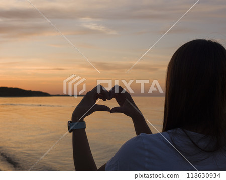 As the sun sets on the seashore, young woman creates a heart with her hands, symbolizing love and peace. Her heart-shaped gesture is captured in a moment of connection with nature's beauty. 118630934