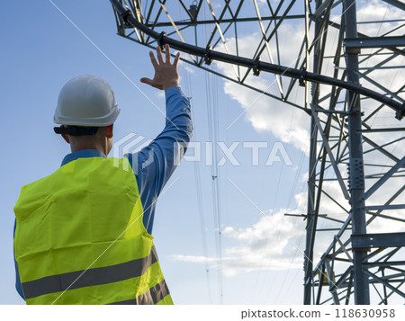 An energy engineer in white helmet and reflector vest gestures towards electric tower See energy engineer's role in maintaining power infrastructure. Energy engineer ensures seamless energy supply. 118630958